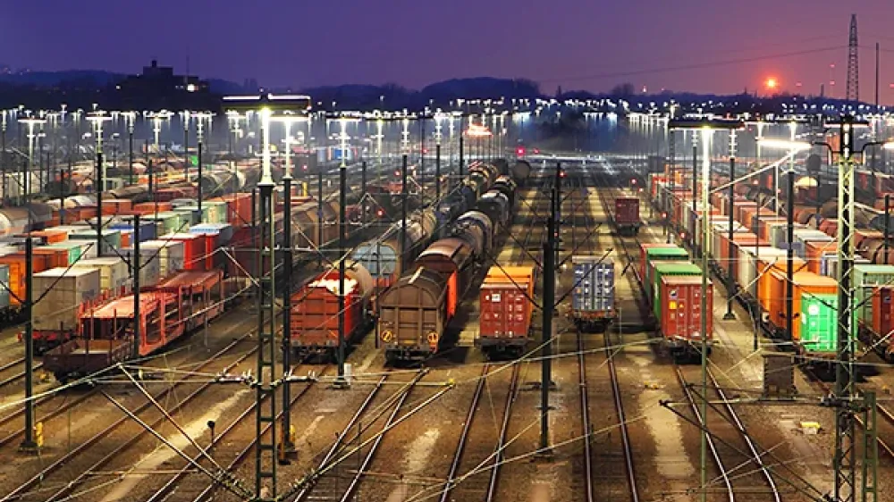 Area ferroviaria con numerosi binari, scambi e linee elettriche sullo sfondo dello skyline cittadino.