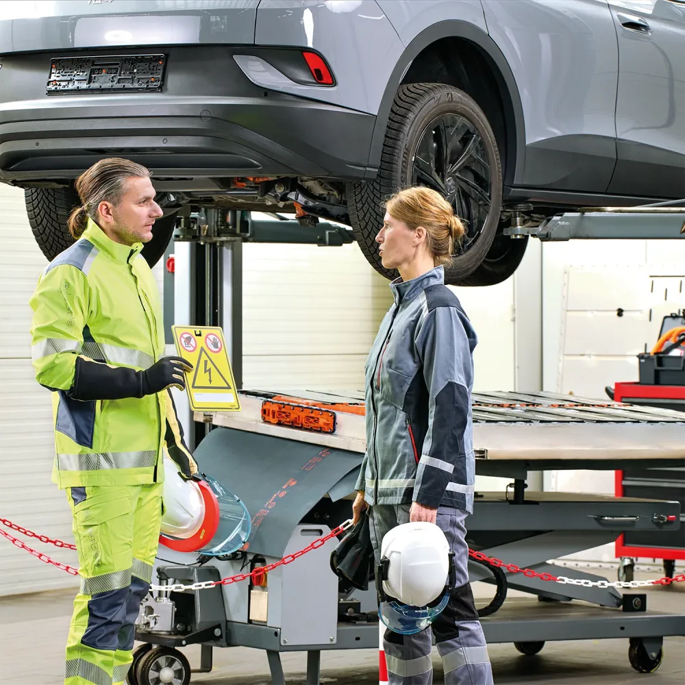 Due operai in tenuta di sicurezza discutono sotto un veicolo elettrico sollevato. Uno tiene in mano un cartello di pericolo. Entrambi indossano caschi e indumenti ad alta visibilità. Nel garage sono visibili attrezzi e macchinari.
