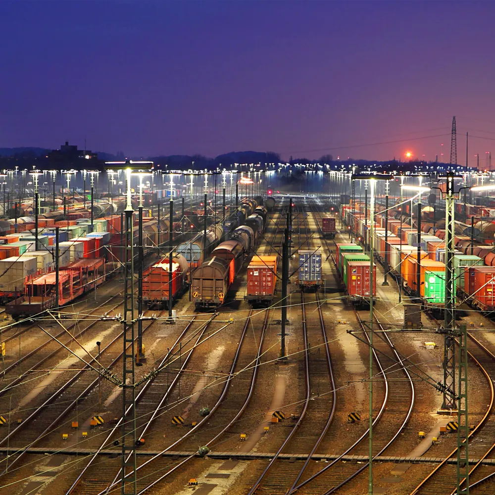 Numerosi treni merci e container sono allineati in uno scalo ferroviario illuminato al tramonto, con i binari che convergono in lontananza e il sole che tramonta all'orizzonte.