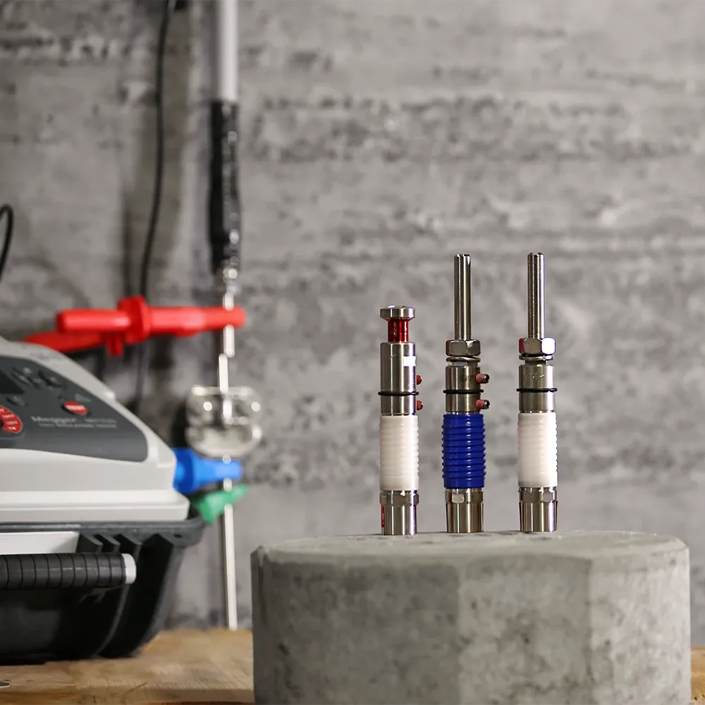 Tre oggetti metallici cilindrici con elementi rossi, blu e bianchi su una superficie in calcestruzzo davanti a un dispositivo con pannello di controllo.