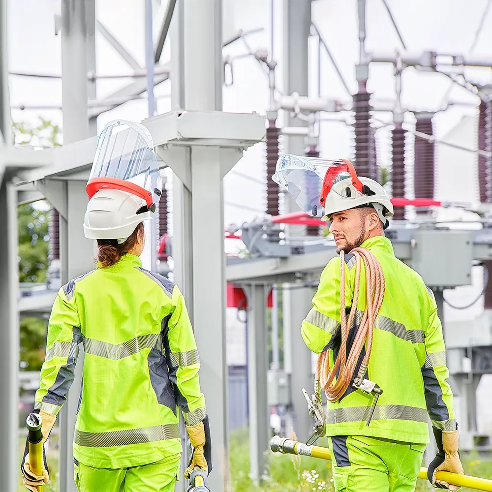 Una donna e un uomo in abbigliamento di protezione completo davanti agli impianti di una sottostazione elettrica.