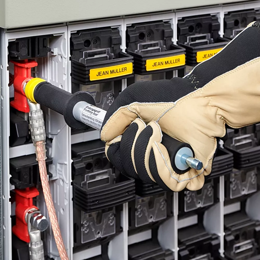 Close-up of a gloved technician connecting a grounding tool to a fuse switch in an electrical distribution cabinet with labeled modules.