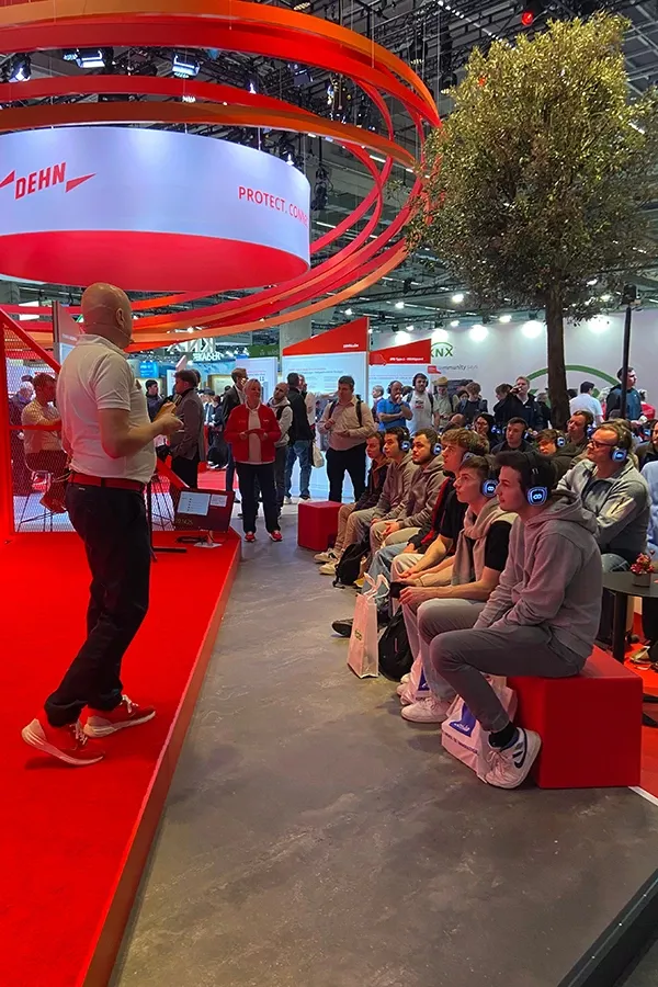 Man with microphone stands on red stage in front of seated audience in modern exhibition hall with red Dehn logo and tree in background