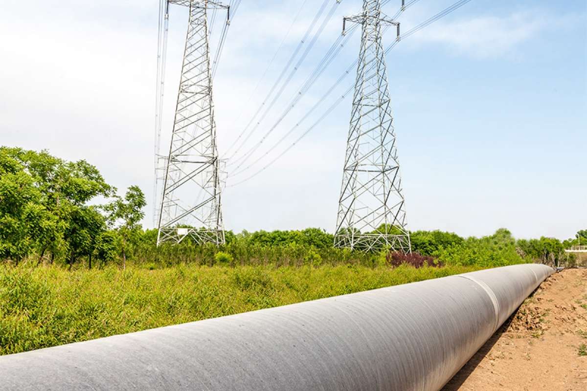 Linea ad alta tensione e conduttura su prato verde con bosco sullo sfondo.